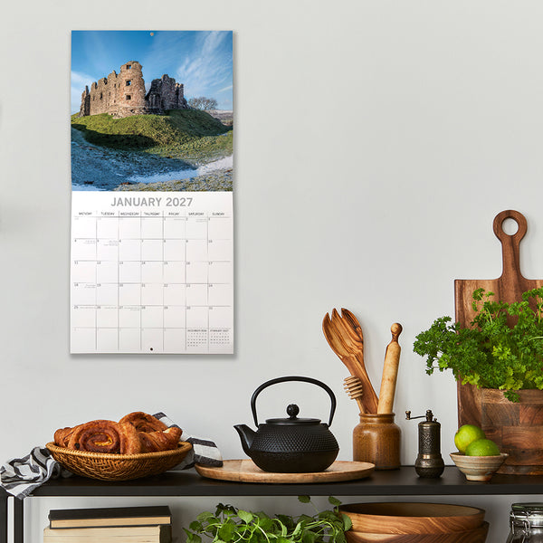 2027 Cumbria Calendar with a scenic landscape on a wall above a kitchen counter with a teapot and croissants.