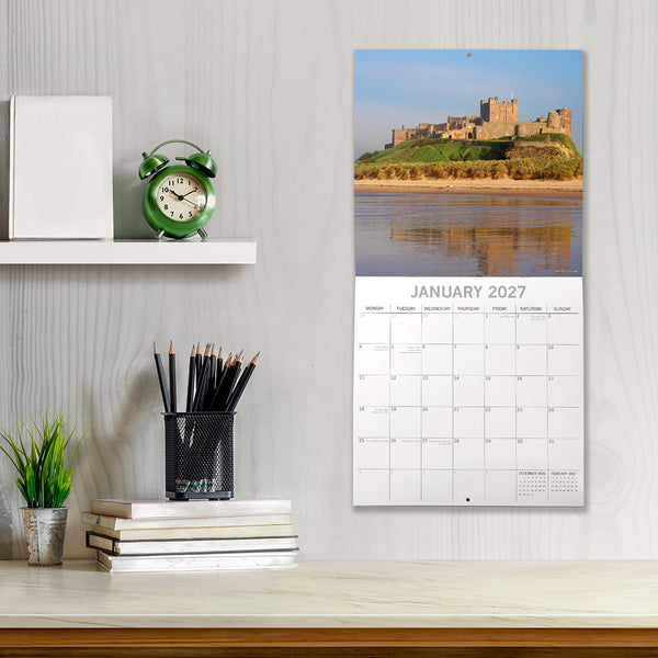 2027 Northumberland Calendar with January 2027 view of Bamburgh castle, placed on a shelf with stationery items.