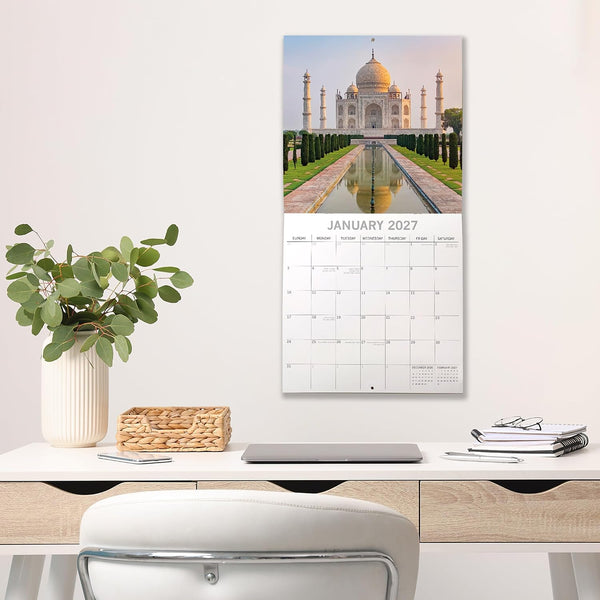 A vertical wall calendar print featuring a photograph of the Taj Mahal above a January 2027 monthly grid, displayed on a light wall above a desk with a plant, laptop, and books.