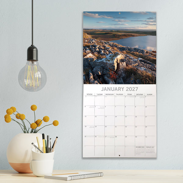 Yorkshire Calendar for January 2027 on a wall next to a light bulb and vase with flowers.