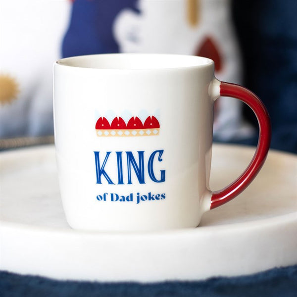 A white ceramic mug with a red handle and 'King of Dad jokes' text, featuring a crown symbol.