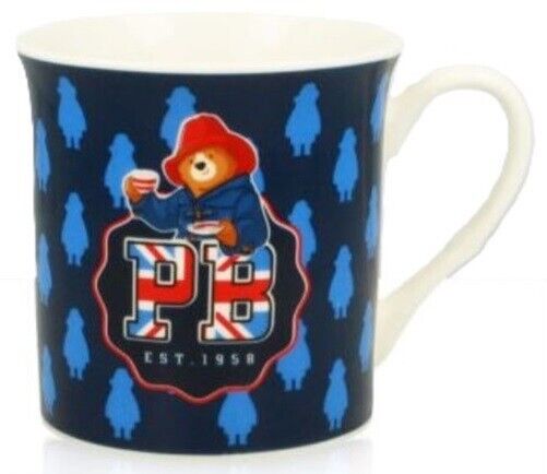 A ceramic mug featuring a colorful design with Paddington Bear, a blue patterned background, and the text 'PB' Est. 1958.